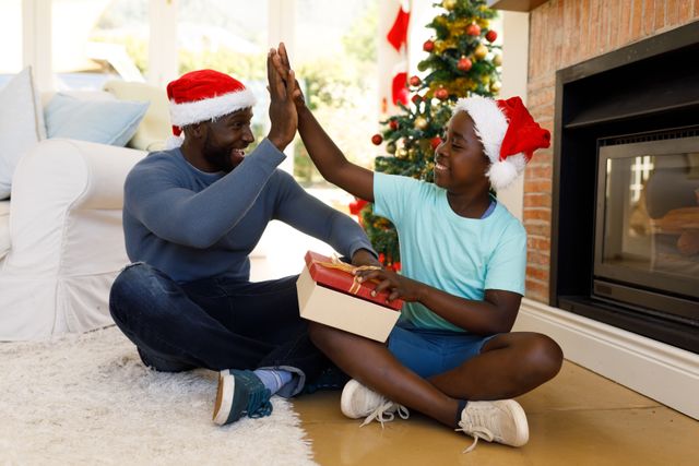 An african american father handing his son his christmas gift while sitting on the floor beside the chimney. the father offers a high five while his soon is holding the gift on his lap.