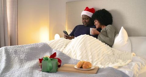 Mid-adult couple relaxing in bed wearing santa hat browsing phone holding mug and gift