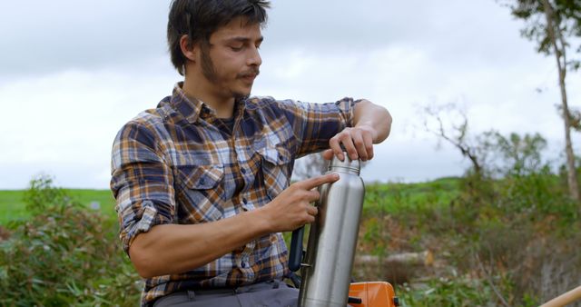 A young Latino man is outdoors, preparing a thermos with a chainsaw and greenery in the background, with copy space. His casual attire and the equipment suggest he might be taking a break from outdoor work or forestry.