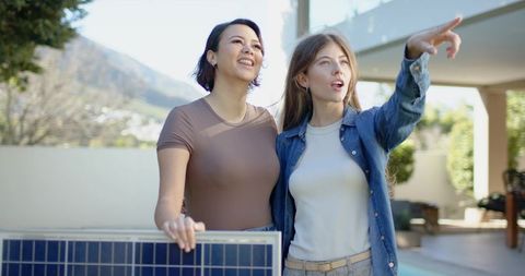 Two women holding portable solar panel and pointing toward hillside from backyard patio