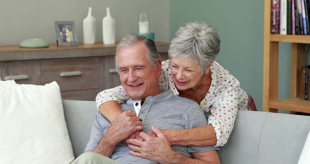 An elderly man and woman sharing a tender moment while seated on a comfortable sofa. This can be used to show love, companionship, and the joy of spending time at home during retirement. Ideal for campaigns on elderly care, retirement living, and family bonds.