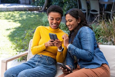 Indian and african-american friends sharing phones and laughing on sunny backyard sofa