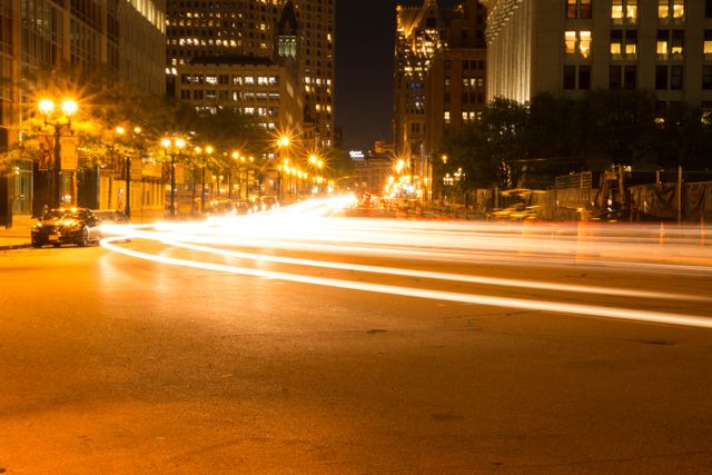 Long exposure captures car light trails on urban street at night. Illuminated buildings and lit lamp posts enhance scene. Useful for projects involving urban nightlife, traffic flow, city energy, nighttime photography.