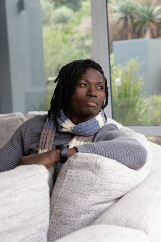 Contemplating african american man sitting on sofa wearing sweater and scarf by window