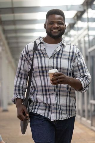 Smiling african american commuter walking with coffee in urban glass corridor plaid shirt