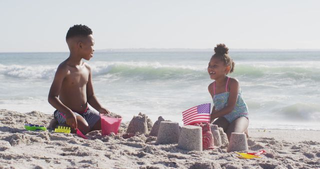 Children building sandcastles on a sunny beach, having fun and enjoying summer activities by the ocean. The scene includes a boy and a girl, highlighting sibling bonding and outdoor play. The American flag adds a patriotic element, ideal for themes related to holidays such as Memorial Day, Independence Day, and Veterans Day. Perfect for uses in family vacation advertisements, summer event promotions, and educational materials on childhood development.