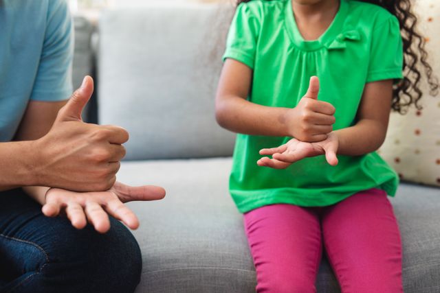 Father and daughter using sign language while sitting on a couch at home. This image can be used for topics related to communication, disability awareness, family bonding, parenting, and educational materials on sign language. It highlights the importance of inclusive communication and family support.