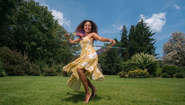 Joyful woman twirling colorful hula hoop in yellow floral sundress on summer lawn