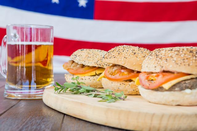Three delicious cheeseburgers with sesame buns, tomatoes, and cheese are placed on a wooden board next to a mug of beer. The background features the USA flag, creating a patriotic and festive atmosphere. Ideal for use in advertisements, social media posts, and articles related to American cuisine, celebrations, barbecues, and national holidays like Independence Day and Memorial Day.