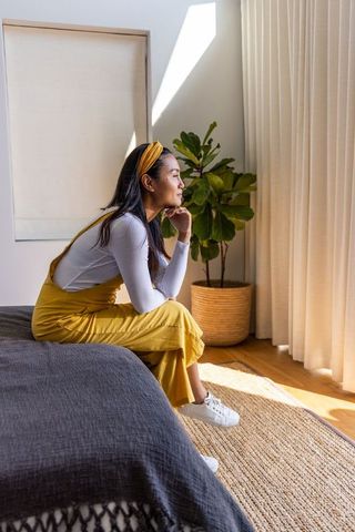 Asian woman in 20s sitting on bed edge wearing mustard pinafore, cozy minimalist bedroom