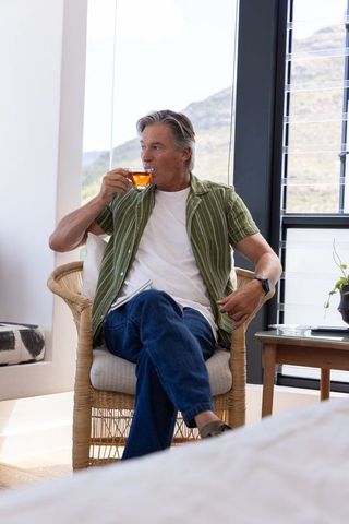 Mature man relaxing in sunlit modern home sipping tea in wicker chair with mountain view