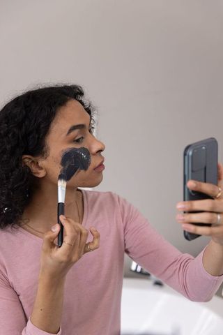 Nonbinary person applying charcoal face mask while checking phone at bathroom mirror
