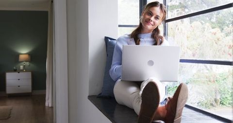 Smiling Woman Using Laptop in Cozy Window Nook at Home