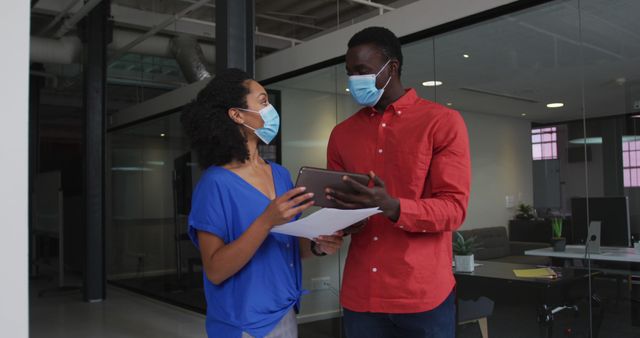 Image shows two office colleagues wearing masks, collaborating using a digital tablet and documents in a modern office space. Useful for articles on workplace collaboration, business practices during COVID-19, teamwork, and the integration of technology in modern professional environments.