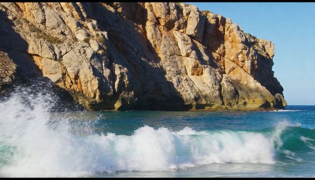 Waves crashing against sunlit rocky cliff with turquoise surf and dramatic coastal headland