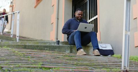 African american man working on laptop while sitting on urban steps with coffee and bag