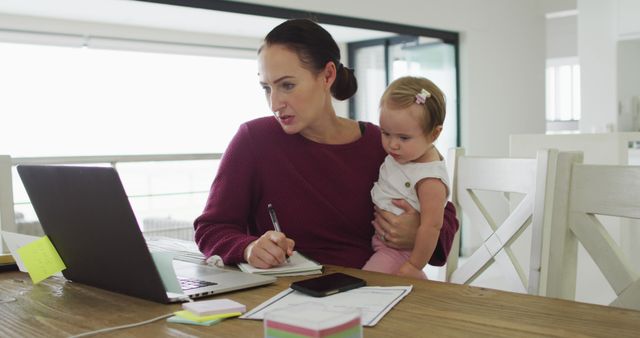 Caucasian mother holding her baby taking notes while working from home - Download Free Stock Photos Pikwizard.com
