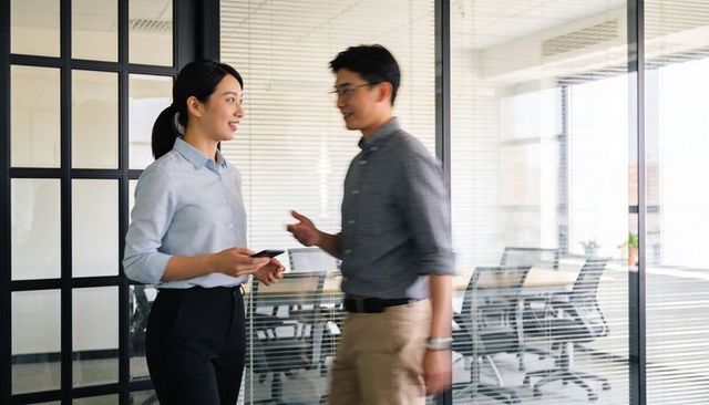 Colleagues discussing project while holding smartphone near glass-walled conference room