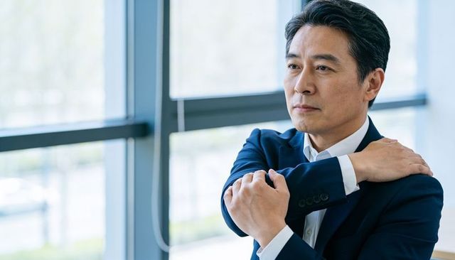 Contemplative asian executive standing by window rubbing shoulder in modern office