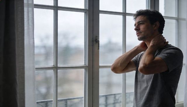 Contemplative man gazing through rain-speckled apartment window during overcast morning