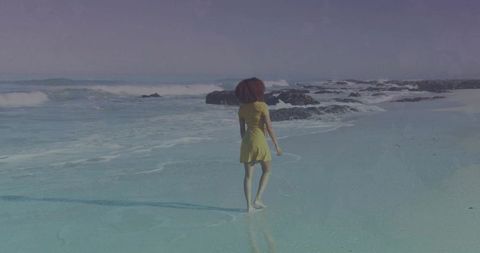 Woman walking barefoot along reflective shoreline in yellow dress with rolling waves