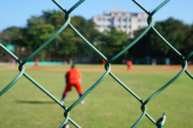 An image of baseball game with players on field seen through green chain link fence. Player in red uniform is visible in foreground with blurred teammates and surroundings in the background. Can be used for articles about sports, team activities, community events in sports facilities, and outdoor sports equipment.