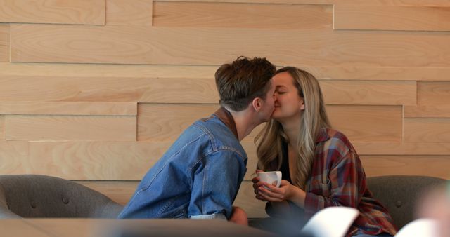 Young couple sharing a romantic kiss in a modern café with stylish wooden panel décor. The couple is casually dressed, with the woman holding a coffee cup. This image is ideal for advertising coffee shops, cafés, dating apps, or romantic relationship concepts.