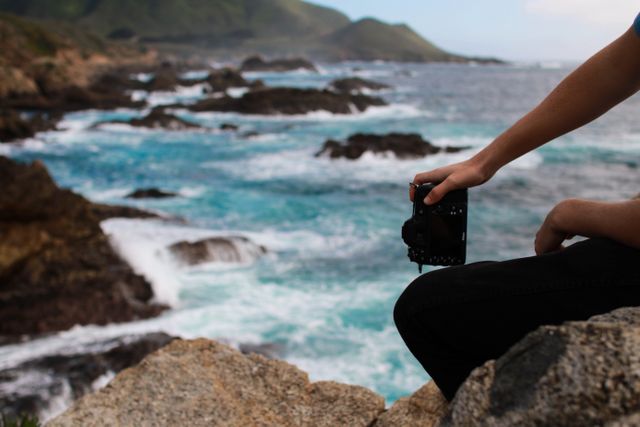 Photographer holding camera, capturing stunning ocean scenery from rocky coast. Waves crashing on shore, landscape appreciator or travel blog, adventure advertisement, outdoor activity promotion.