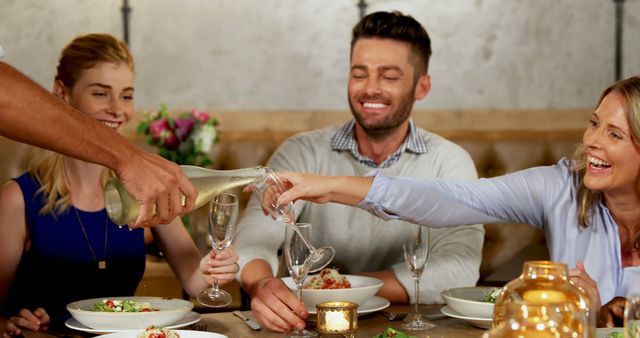Group of friends sitting at a dining table, talking and laughing while wine is being poured into glasses. They are enjoying a meal in a warm, inviting restaurant atmosphere. Perfect for depicting social gatherings, celebrations, and the joy of sharing meals with friends.