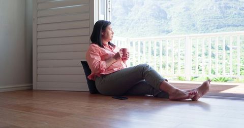 Mature asian woman relaxing on wood floor by balcony door holding mug enjoying mountain view