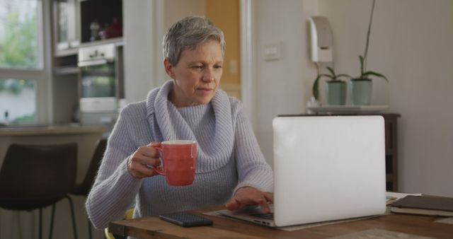 Senior woman working on laptop while drinking coffee in cozy kitchen - Download Free Stock Images Pikwizard.com