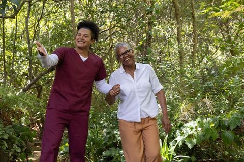 Young caregiver guiding african american senior woman on shaded woodland trail