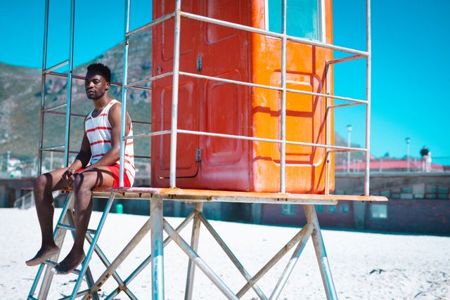 African american young man contemplating while sitting on lifeguard hut on sunny day, copy space. summer, beach, protection, unaltered, lifestyle, enjoyment and holiday concept.