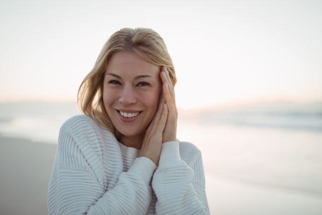 Young woman smiling at beach during dusk, wearing casual sweater. Ideal for use in lifestyle, travel, and wellness content. Perfect for promoting relaxation, happiness, and outdoor activities.