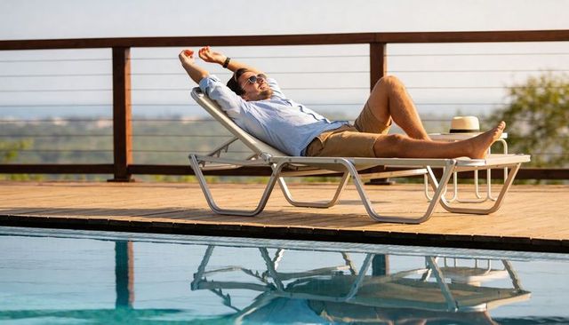 Man relaxing on poolside lounge chair stretching arms in sunlit outdoor deck scene