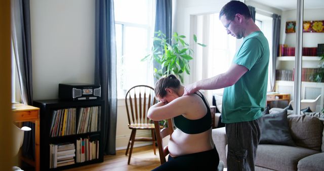 Partner giving massage to pregnant woman in living room. Perfect for content about prenatal care and support, relationships, family health, pregnancy wellness, and home support. Could be used for articles and campaigns on family and maternity services, as well as health and wellness blogs.
