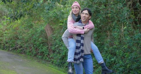 Young couple enjoying piggyback hike on mossy woodland trail in cozy winter clothing