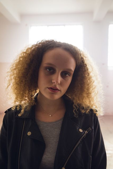 Young Caucasian woman with curly hair standing in an empty warehouse, backlit by sunlight from a window. She is wearing a leather jacket and a casual top, looking straight at the camera with a serious expression. Ideal for use in fashion editorials, urban lifestyle blogs, or advertisements focusing on youth culture and contemporary fashion.