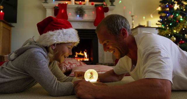 Father and daughter lying on the floor, enjoying Christmas lights in a cozy, festive room. The fireplace glows warmly in the background and stockings hang above. Both are smiling and engaged in a joyful moment, the young girl wearing a Santa hat. Ideal for depicting family bonding, holiday spirit, and Christmas celebrations.
