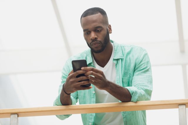 Young African-American man standing indoors, using smartphone while leaning on wooden rail. Suitable for concepts such as communication, technology, modern lifestyle, and social media engagement. Can be used in advertisements, articles, or promotional materials related to mobile technology and social interactions.
