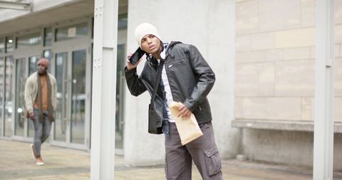 Urban young man listening on phone while holding parcel outside storefront with passerby
