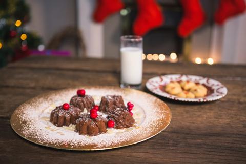 Christmas desserts with red berries and powdered sugar on a golden plate, accompanied by a glass of milk. In the background, festive decorations and Christmas stockings are visible. Ideal for holiday-themed promotions, festive recipe blogs, and seasonal greeting cards.