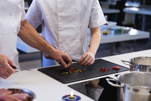 Mid section of two male chefs cooking in a modern busy kitchen, slicing chilli peppers on chopping board. Cookery class at a restaurant kitchen. Workshop cooking food.