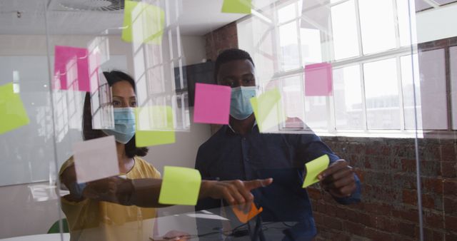 Diverse male and female business colleagues wearing face masks putting sticky note on glass wall. working in a modern office during covid 19 coronavirus pandemic.