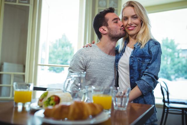 Couple enjoying a romantic moment in a cafe, with breakfast items like juice and croissants on the table. Ideal for use in advertisements for cafes, dating services, or lifestyle blogs focusing on relationships and romance.