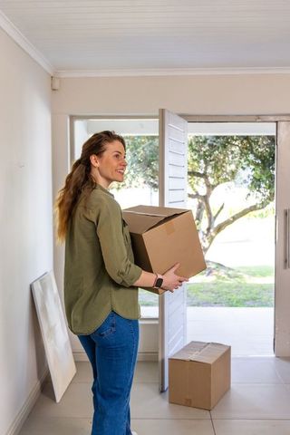 Young woman carrying moving box and smiling in sunlit suburban home entryway