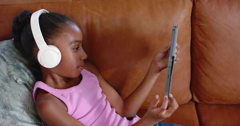 Child reclining on leather couch using tablet with white headphones enjoying cozy screen time