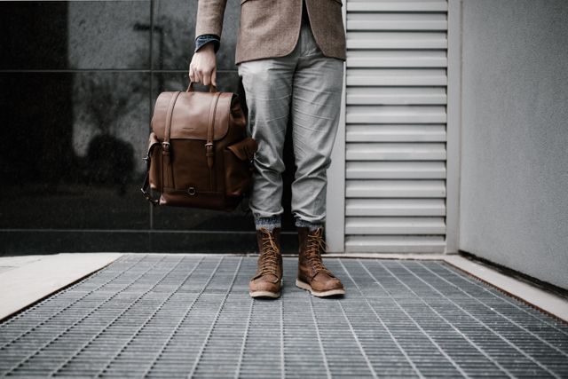 Businessman holding a brown leather backpack and wearing casual yet stylish clothes including trousers and brown boots. Might be used for themes such as urban commuting, professional lifestyle, modern business fashion, or street style. Ideal for blogs, advertisements, or fashion magazines.