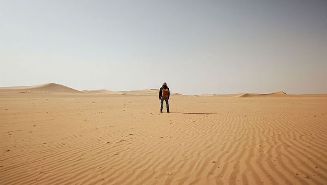 Solitary traveler walking across vast sunlit desert dunes with rippled sand and long shadow