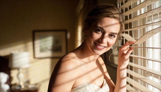 Smiling woman peeking through blinds at golden hour holding blind slat in strapless gown